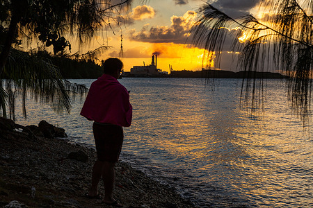 A man watches the sunset from the shoreline of Piti.  In the background, Cabras Power Plant emits smoke in the distance. The plant is being modernized following years of using older technology which generated considerable pollution. While construction delays and other issues risk pushing completion of the project to 2025 and beyond, The improvements will dramatically reduce air pollution and the Guam Power Authority will pay a one-time $400,000 civil penalty rather than face hundreds of millions of dollars in fines for continuing to violate Environmental Protection Agency rules. Disclaimer: This image was captured during the global response to the COVID-19 pandemic. The contents of this image reflect the guidance communicated by local public health authorities at the time of its capture. Please note, public health guidance differs among countries and is indicative of the local context.